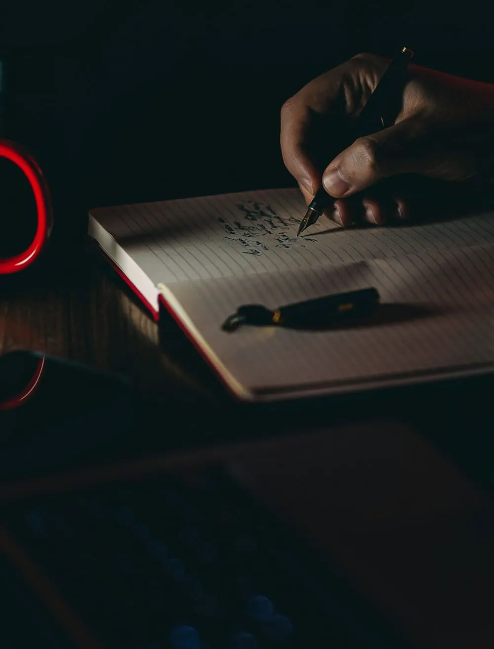 A hand writing in a notebook by lamplight, a pen resting on the open page.