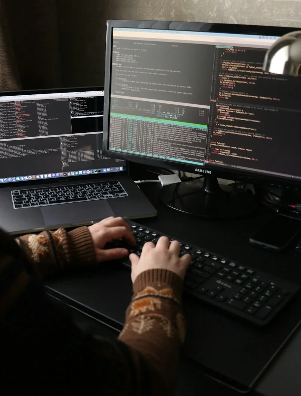 Hands at a keyboard in front of two screens of code, in a darkened room.
