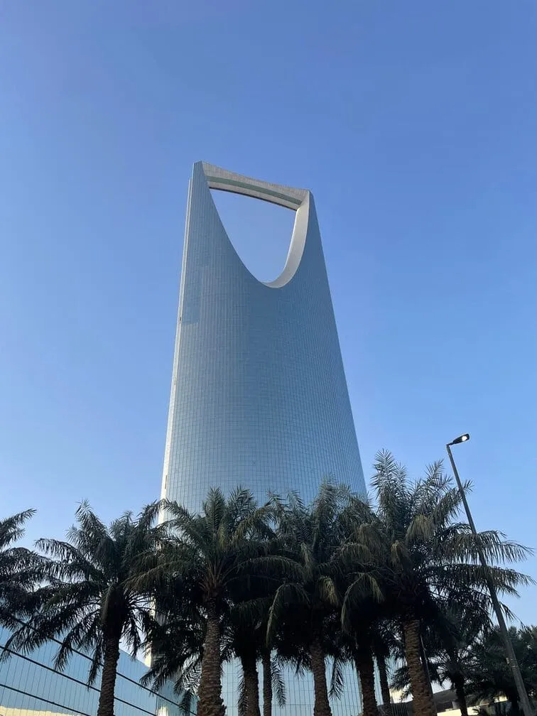 The Kingdom Centre in Riyadh seen from below, a row of palms at its foot and a clear sky above.