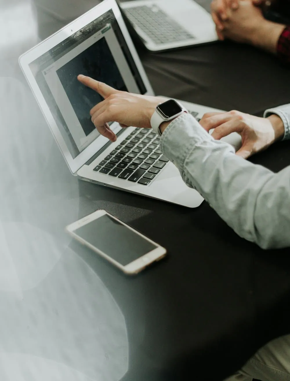 Two people at a desk, one pointing at a laptop screen, a phone and a tablet beside it.